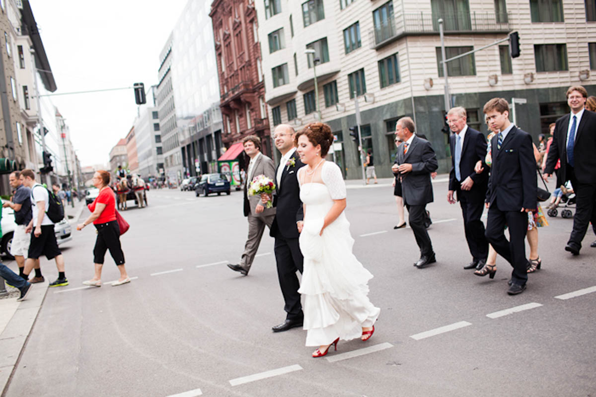Braut im Brautkleid läuft mit ihren Gästen lachend über eine belebte Straße