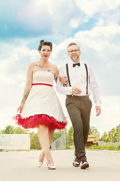 Rockabilly-Brautpaar: Braut im kurzen Spitzenkleid mit rotem Petticoat und Bräutigam mit Hosenträgern und Fliege