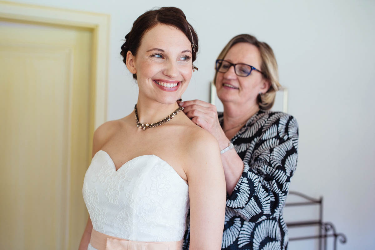 Mutter legt der lachenden Braut im trägerlosen Brautkleid die Kette um