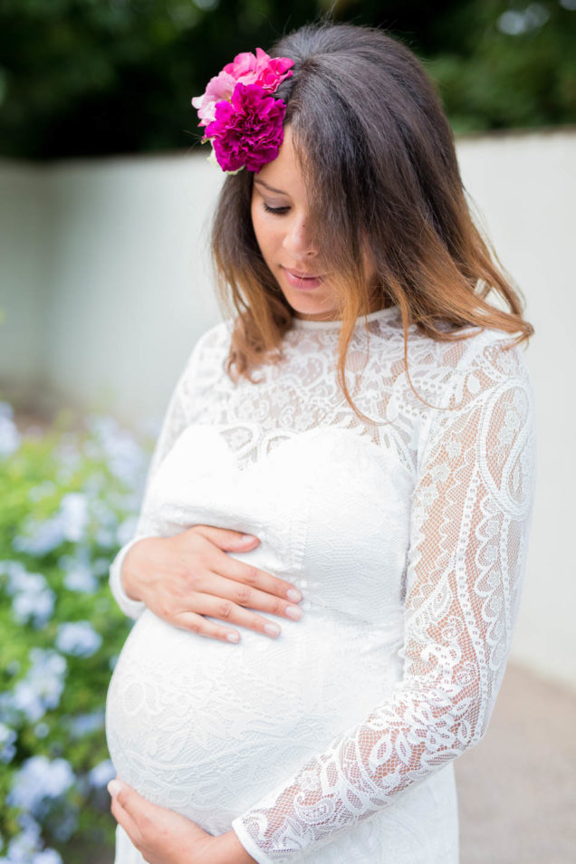 Schwangere Braut im Spitzenkleid mit langen Ärmeln hält ihren Babybauch, blaue Blüten im Hintergrund