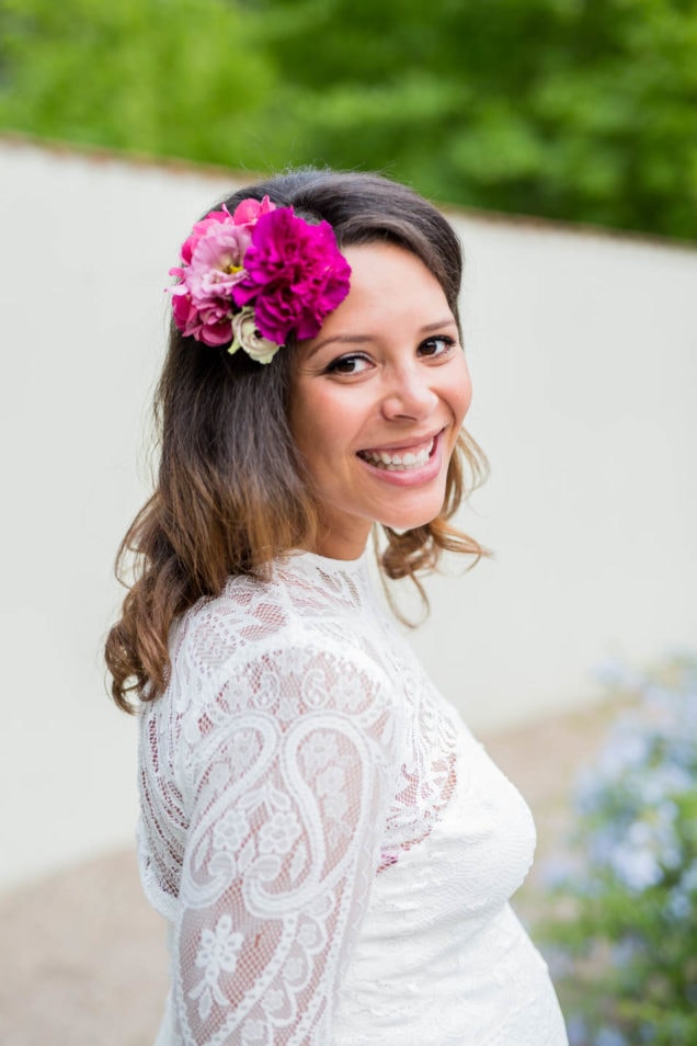 Lachende Braut mit pinkem Blumenschmuck im Haar und hochgeschlossener Spitze