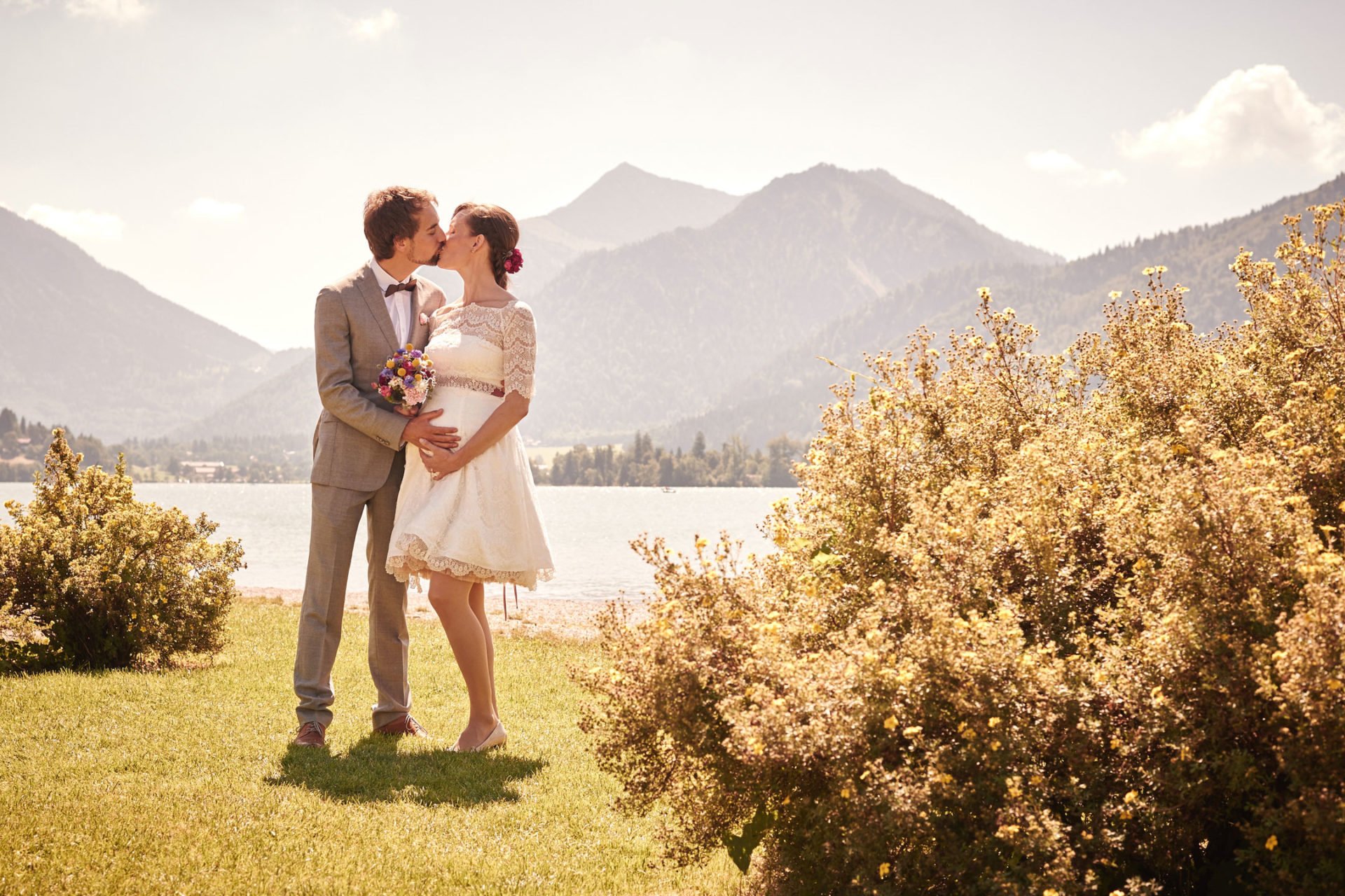 Umstandsbrautkleid kurz – schwangere Braut küsst ihren Mann im kurzen Brautkleid am See