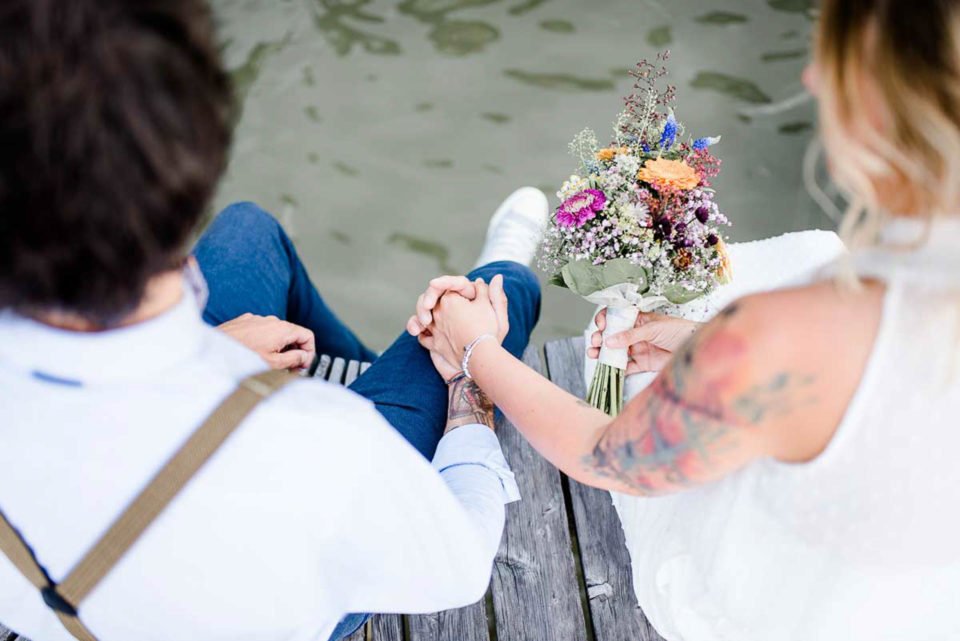 Freie Trauung am See: Brautpaar sitzt am Steg und hält Händchen 