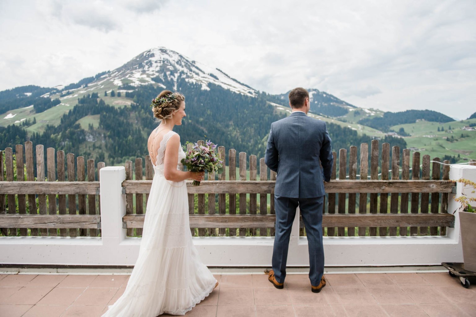 Braut im Boho-Kleid geht auf einer Terrasse auf den Bräutigam zu, der mit dem Rücken zu ihr auf die Berge blickt