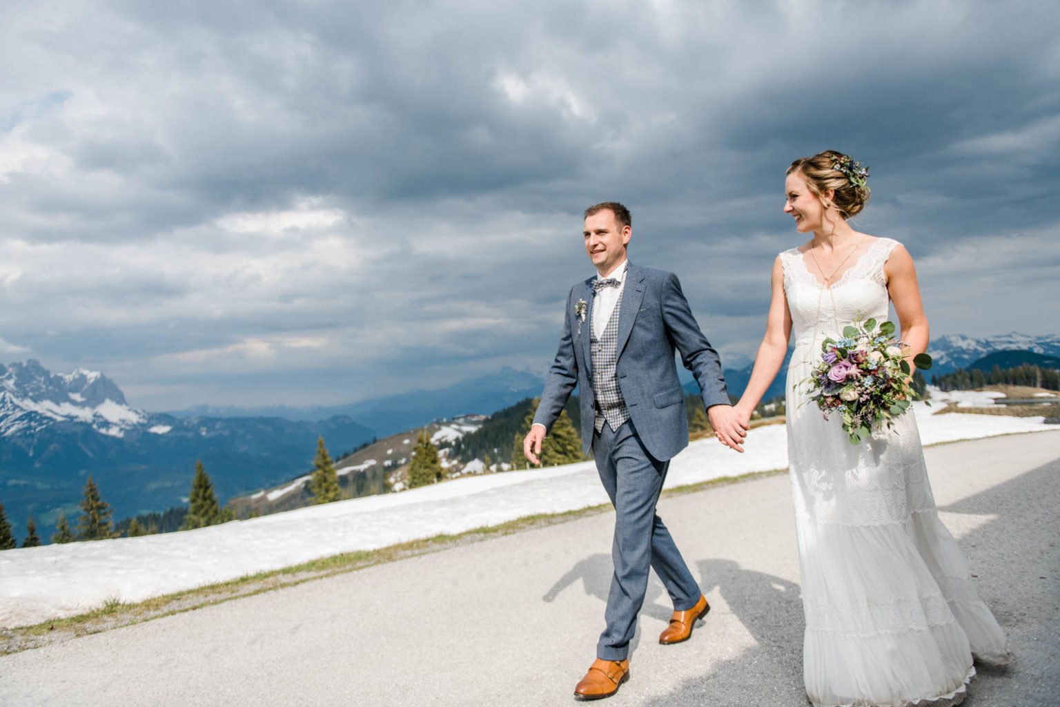 Brautpaar geht Hand in Hand einen Weg entlang, dahinter Berge und Wolken