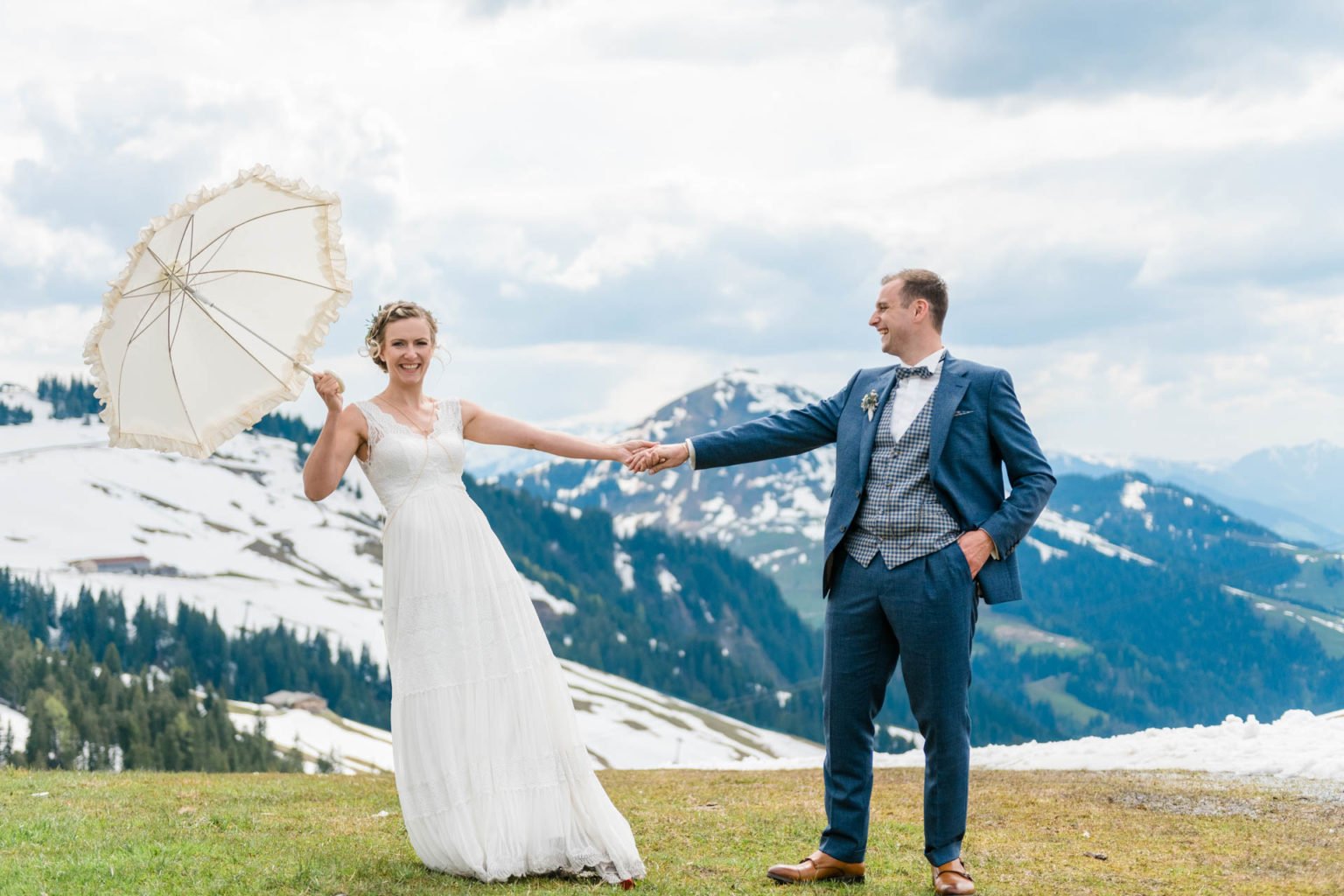Braut mit weißem Sonnenschirm reicht dem Bräutigam die Hand, im Hintergrund Schneefelder