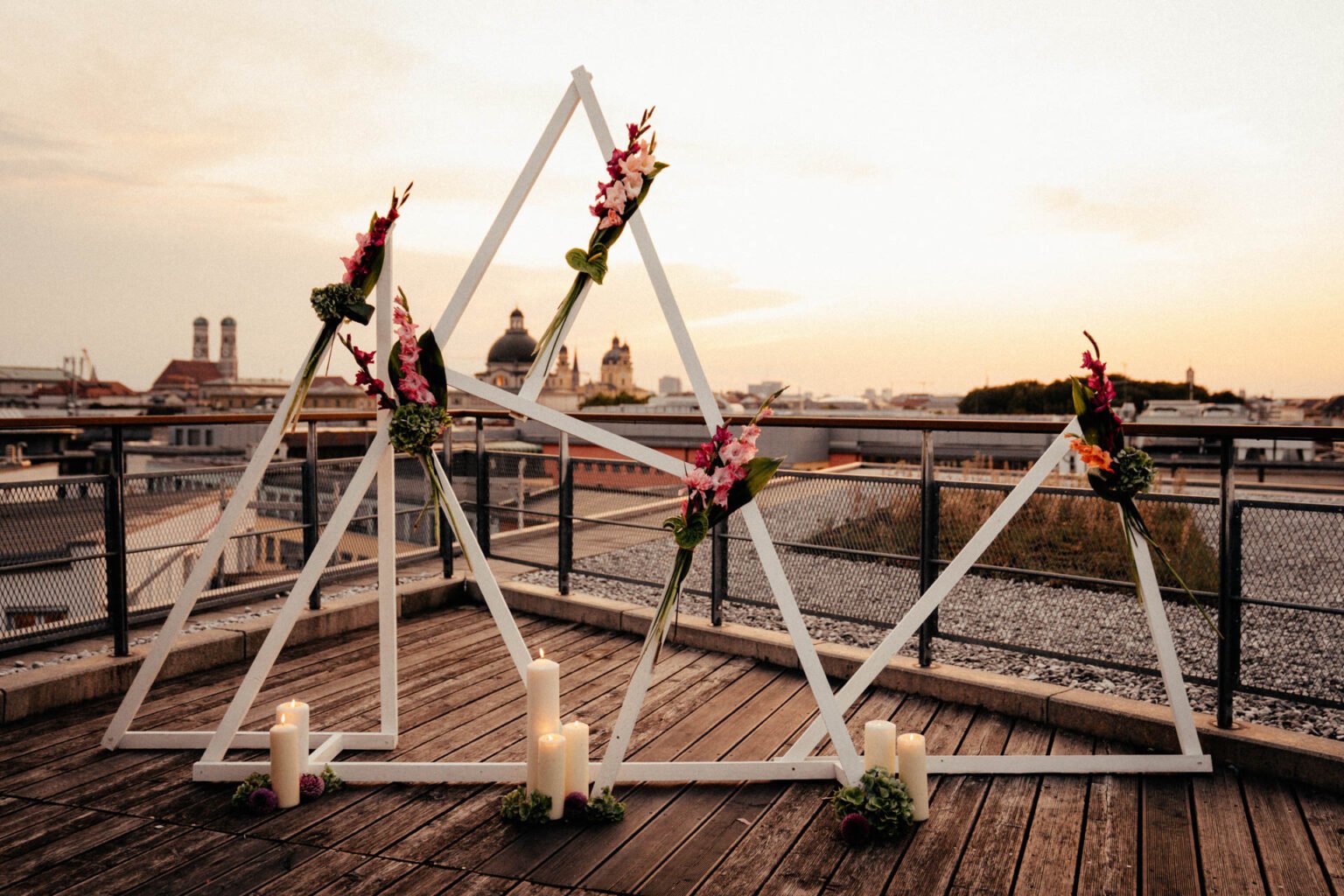 Geometrischer Traubogen aus weißen Holzdreiecken mit pinken Blüten auf einer Dachterrasse über München