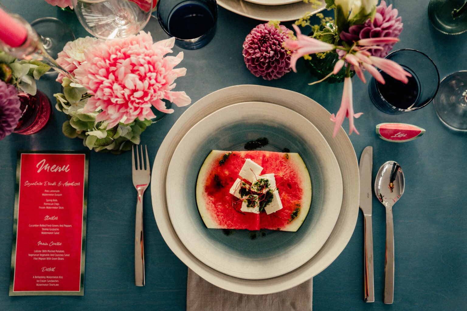 Gedeck mit Melonenscheibe, Menükarte in Pink und Dahlien auf blauem Tisch