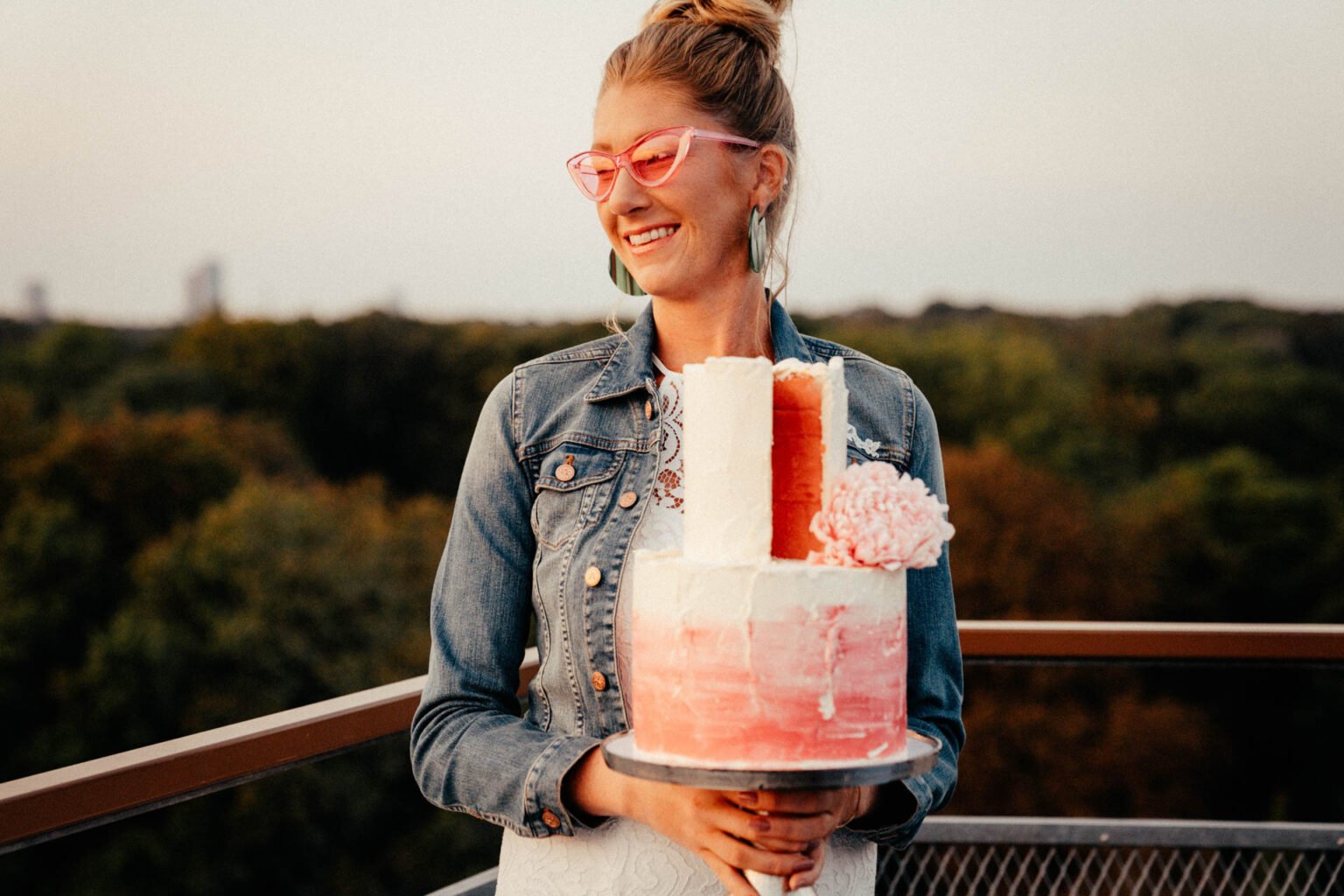 Braut mit Jeansjacke und pinker Sonnenbrille hält lachend die Hochzeitstorte