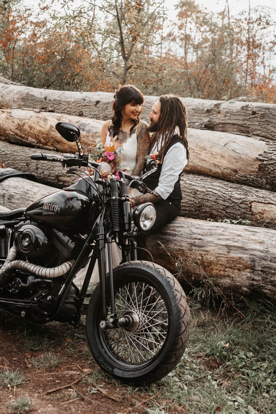 Braut mit Tattoos sitzt neben Bräutigam auf Holzstämmen hinter Harley
