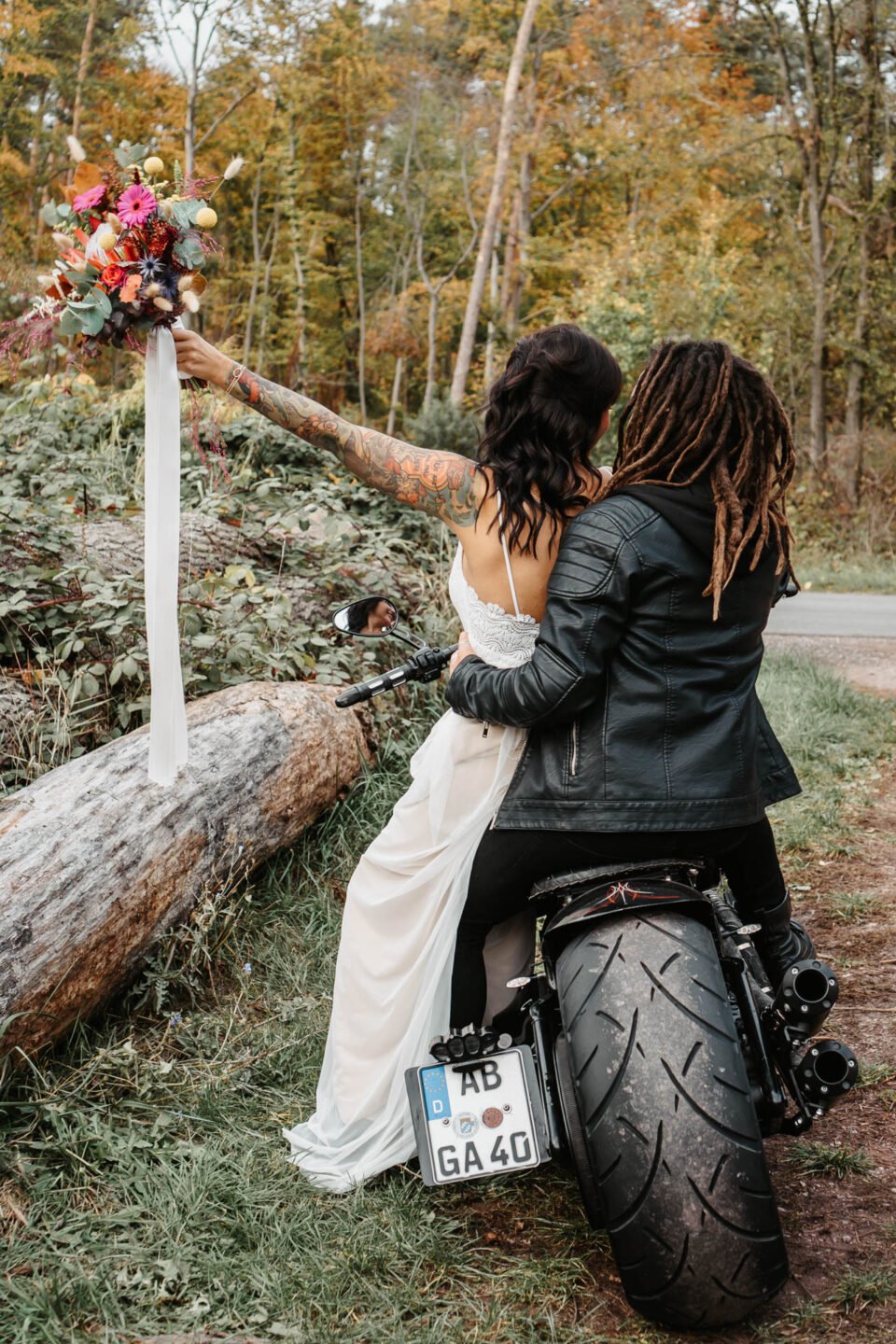tätowierte Braut von hinten als den Arm mit Brautstrauß von sich streckt und neben Bräutigam mit Lederjacke auf Motorrad sitzt 