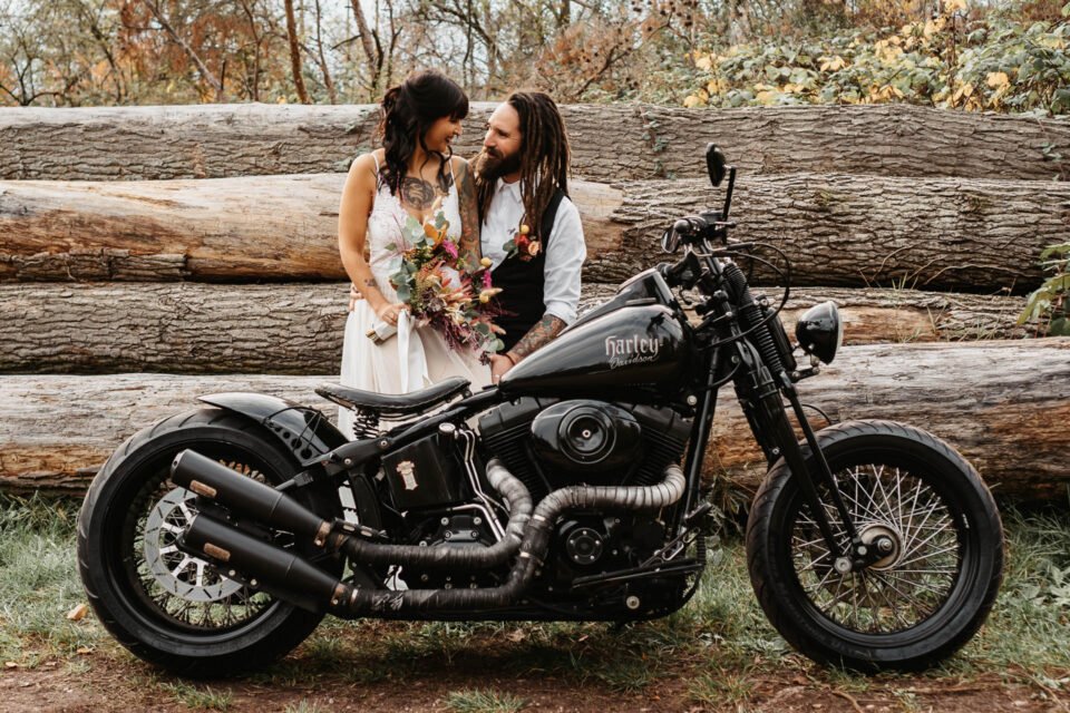 Brautpaar mit Tattoos steht hinter Harley und lächelt sich an