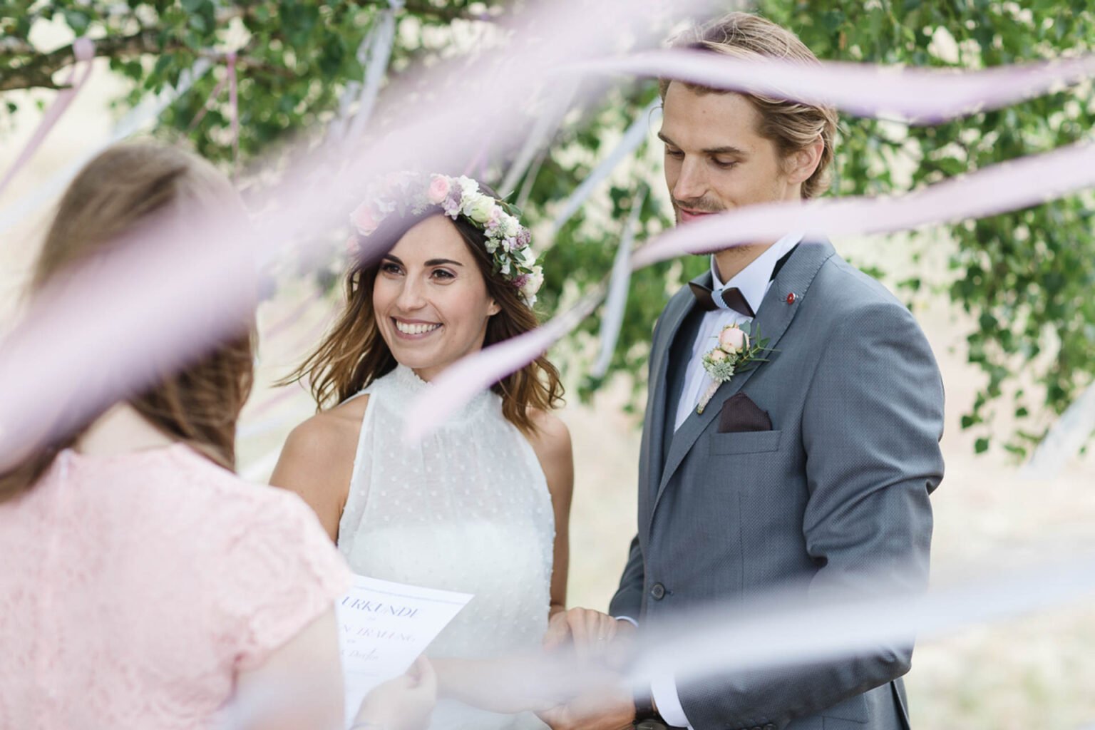 Braut mit Blumenkranz und Bräutigam im grauen Anzug lächeln sich unter Birken an, im Vordergrund rosa Bänder