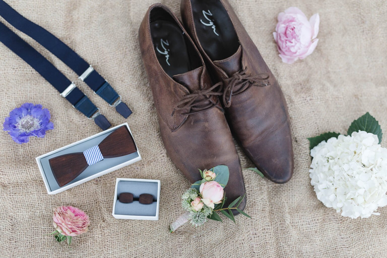 Braune Lederschuhe, blaue Hosenträger, eine Holzfliege und Blüten auf einer Juteunterlage