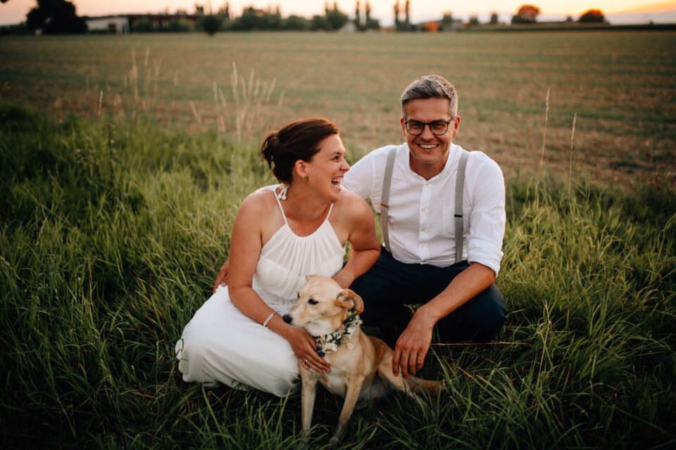 lachendes Brautpaar mit Hund auf Wiese bei Gartenhochzeit