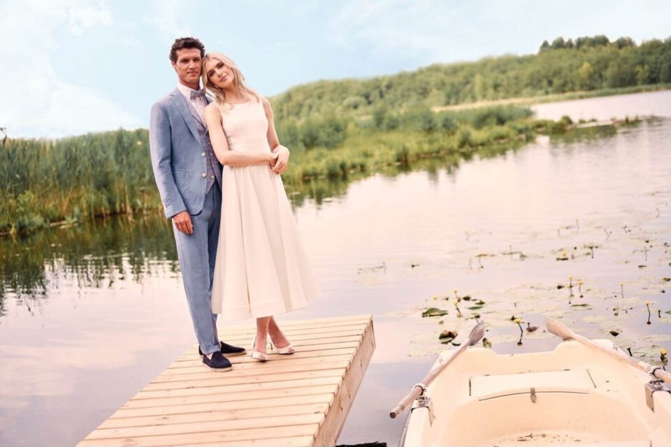 Braut im köchellangen Brautkleid auf einem Stag am See vor Bräutigam im batikblauen Anzug.