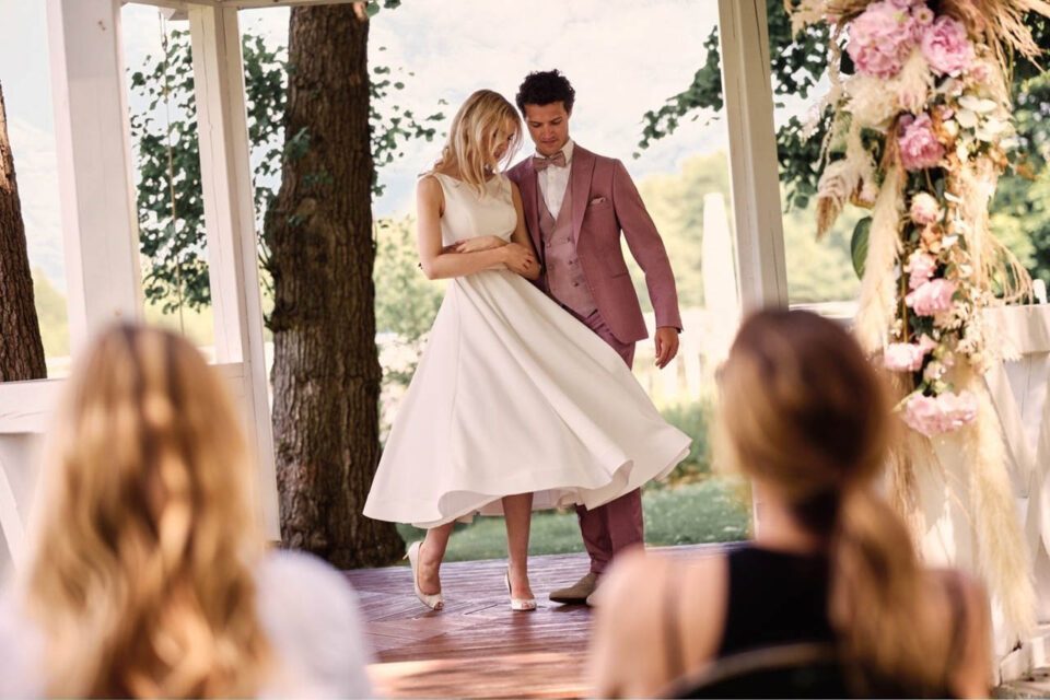 Braut mit köchellangem Brautkleid tanzt mit Bräutigam im Roséfarbenen Anzug