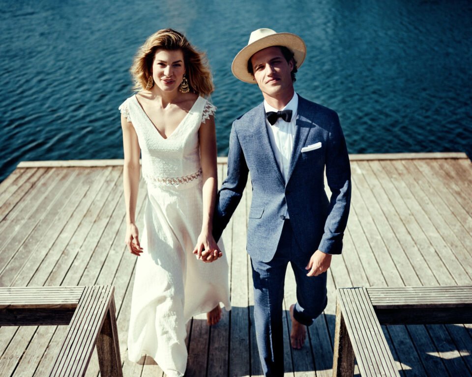 Brautpaar am Wasser. Braut trägt zweiteiliges Brautkleid in Bio Baumwolle und Bräutigam einen dunkleblauen Vintage Anzug