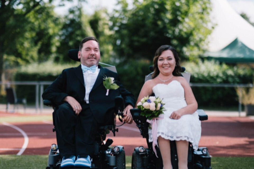 Hochzeit im Rollstuhl, glückliches Brautpaar hält Händchen