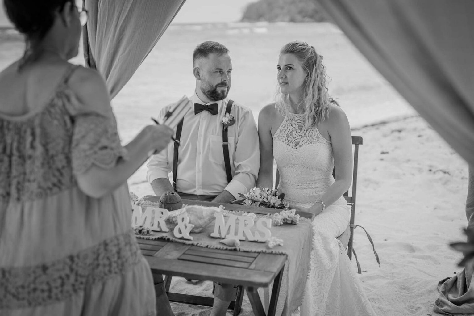 Schwarz-Weiß-Foto: Brautpaar sitzt bei der Trauzeremonie am Tisch, davor die Frau, die die Zeremonie leitet