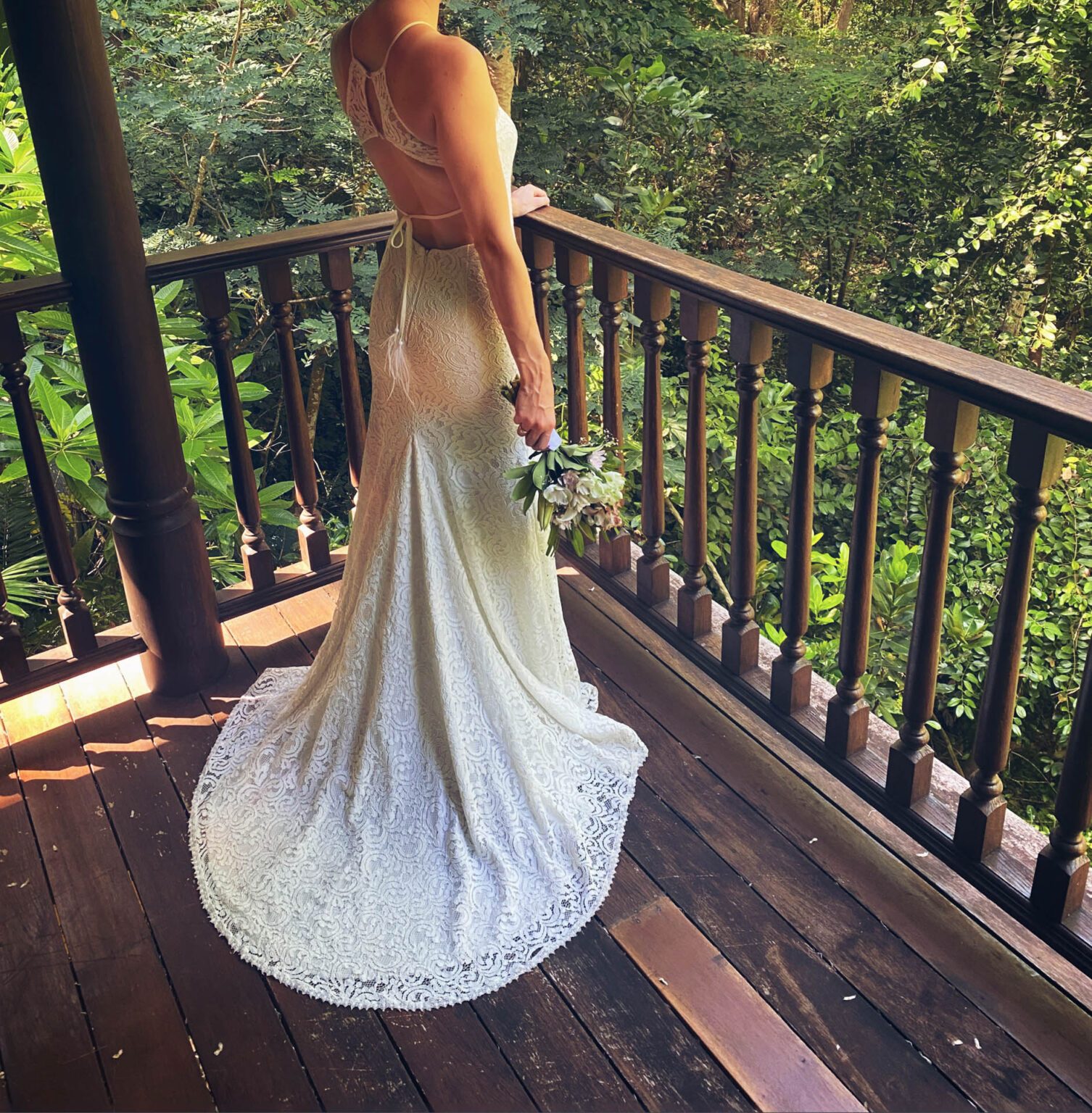 Braut im rückenfreien Spitzenkleid mit Schleppe steht auf einer Holzveranda