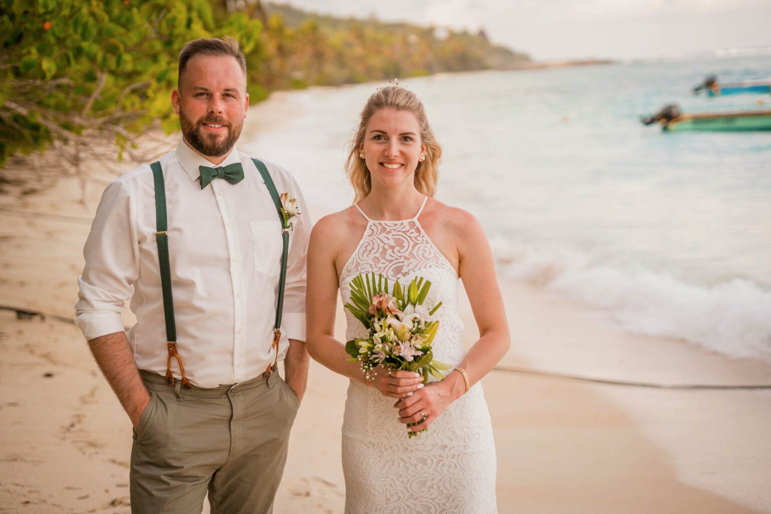 Brautpaar steht lachend am Strand von Mahé, er mit grünen Hosenträgern und Fliege, sie im Spitzenkleid mit Neckholder