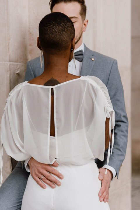 Kurzhaarige, dunkle Braut in Bluse von hinten fotografiert, Bräutigam legt Hände um sie