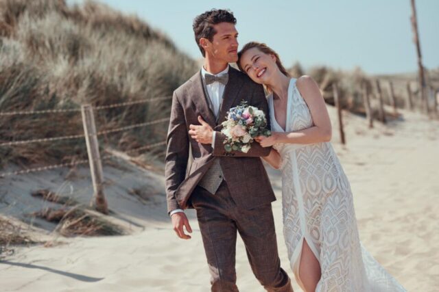Braut im ausgefallenen Brautkleid mit Beinschlitz schmiegt sich in den Dünen an Bräutigam im Vintage Anzug.