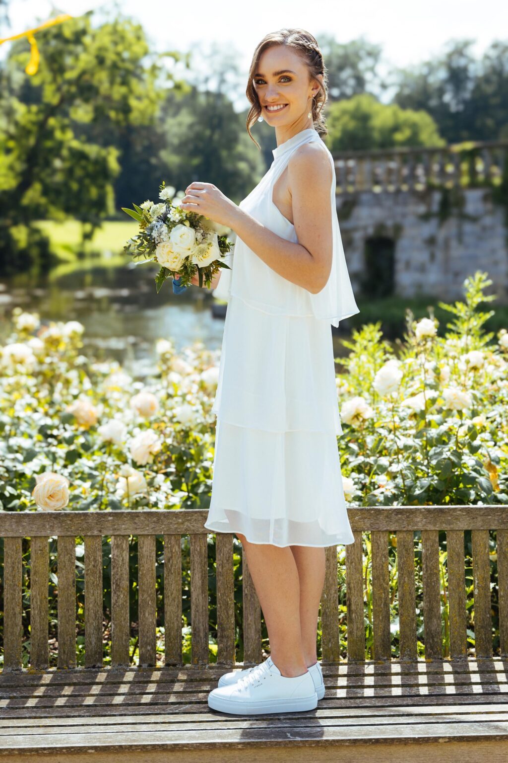 Braut im kurzen Stufenkleid aus Chiffon mit Sneakern auf einer Gartenbank