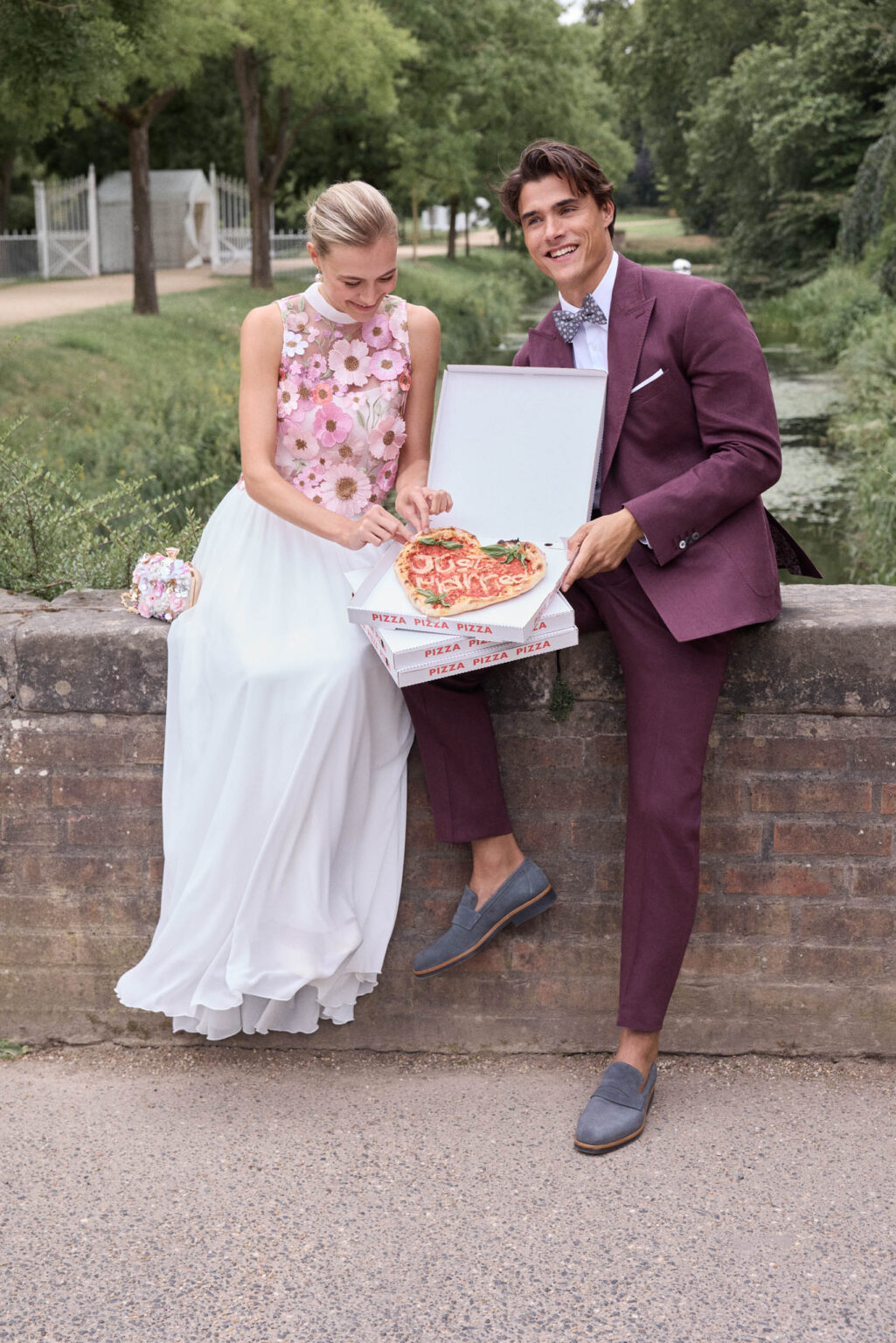 Brautpaar mit Pizza auf einer Mauer, er im beerenfarbenen Anzug, sie im Blumentop