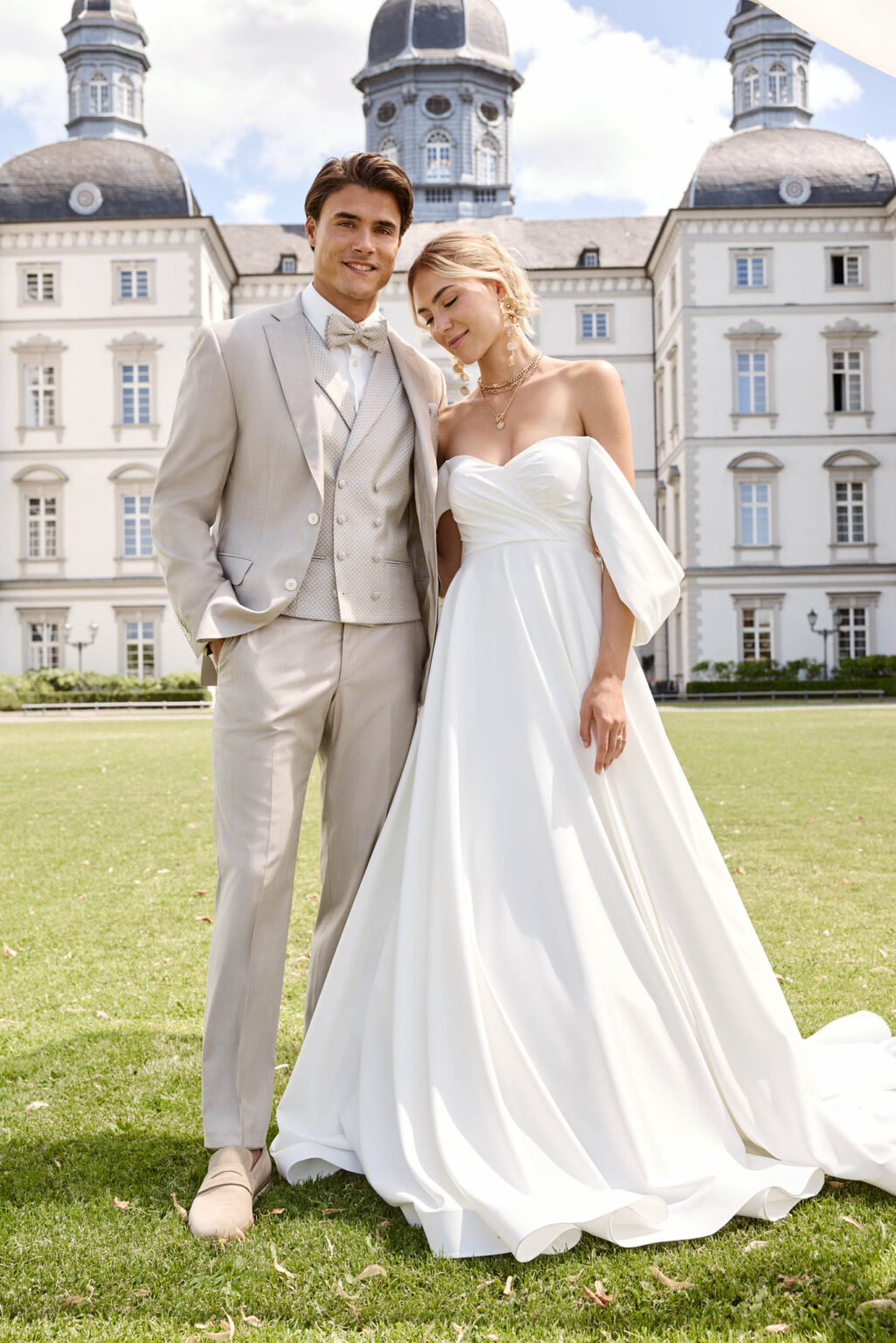Bräutigam im sandfarbenen Dreiteiler und Braut im Off-Shoulder-Kleid vor einem Schloss