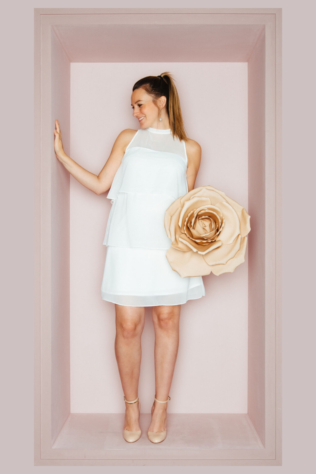 Schwangere Braut in einem kurzen, gestuften Chiffonkleid mit transparentem Stehkragen, daneben eine große Papierrose