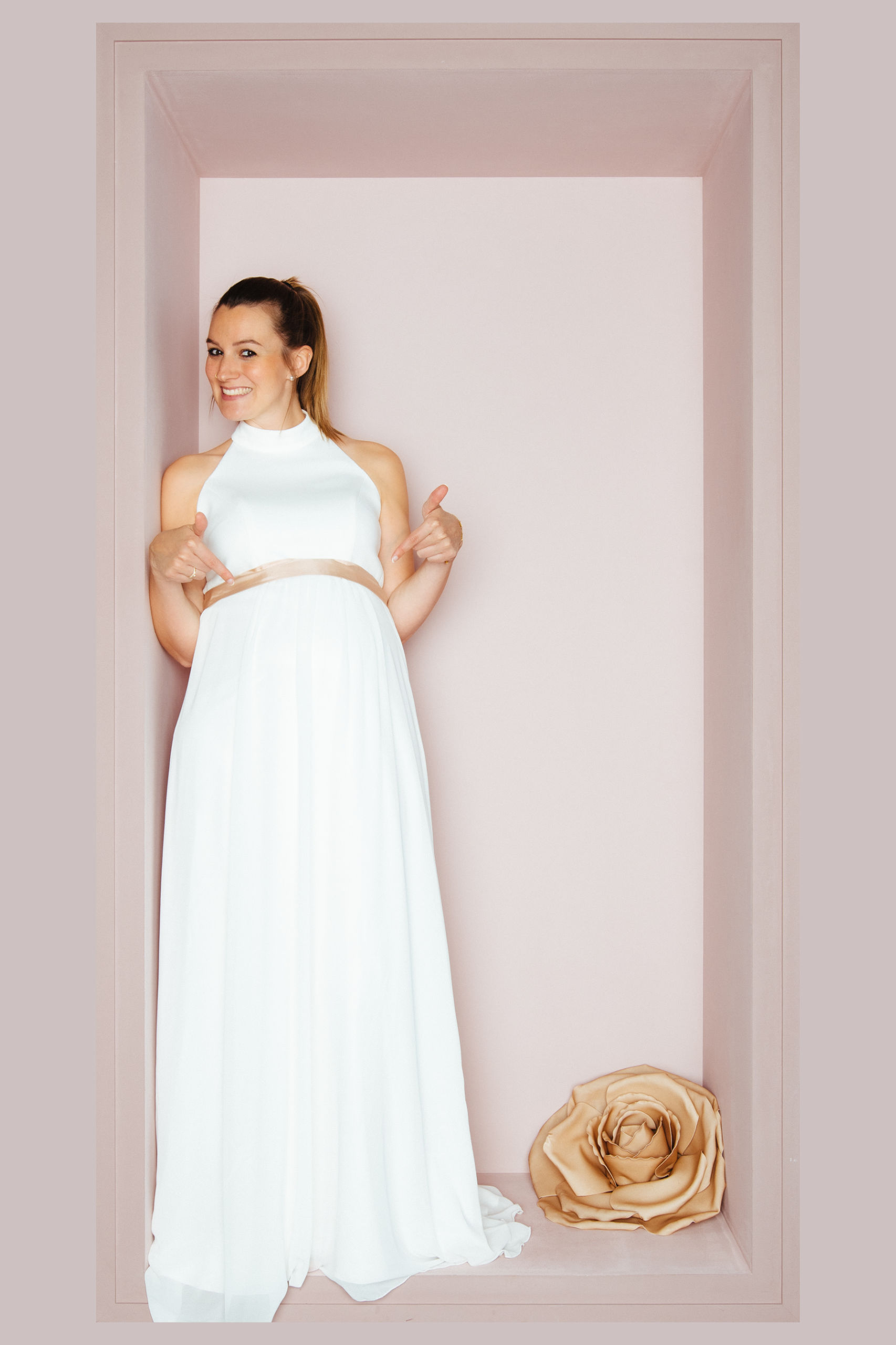 Schwangere Braut in weißem Halterneck-Brautkleid mit beigem Seidenband über dem Babybauch von KUESSdieBRAUT