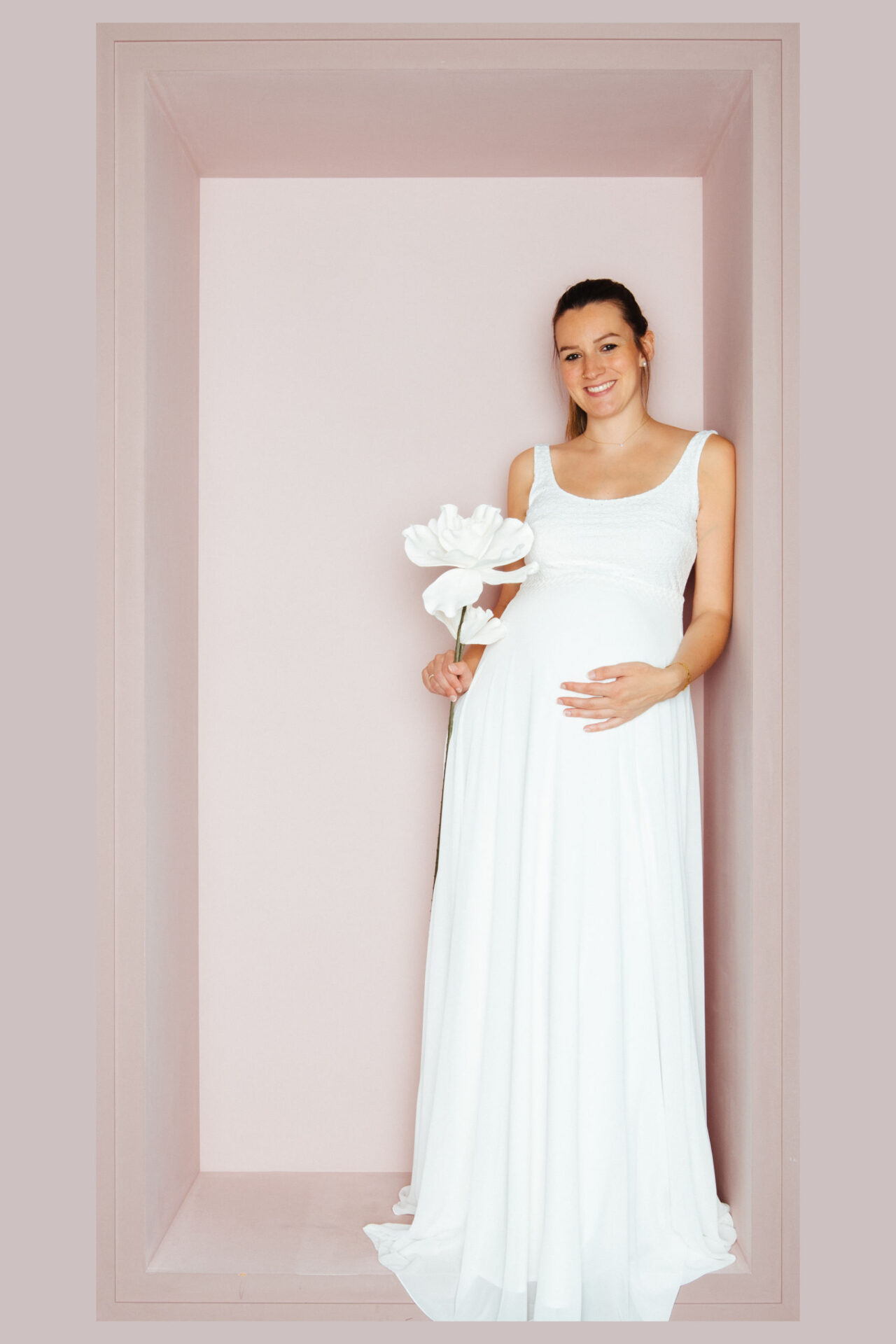 Schwangere Braut im Chiffon-Brautkleid mit breiten Trägern und rundem Ausschnitt, in der Hand eine weiße Papierblume