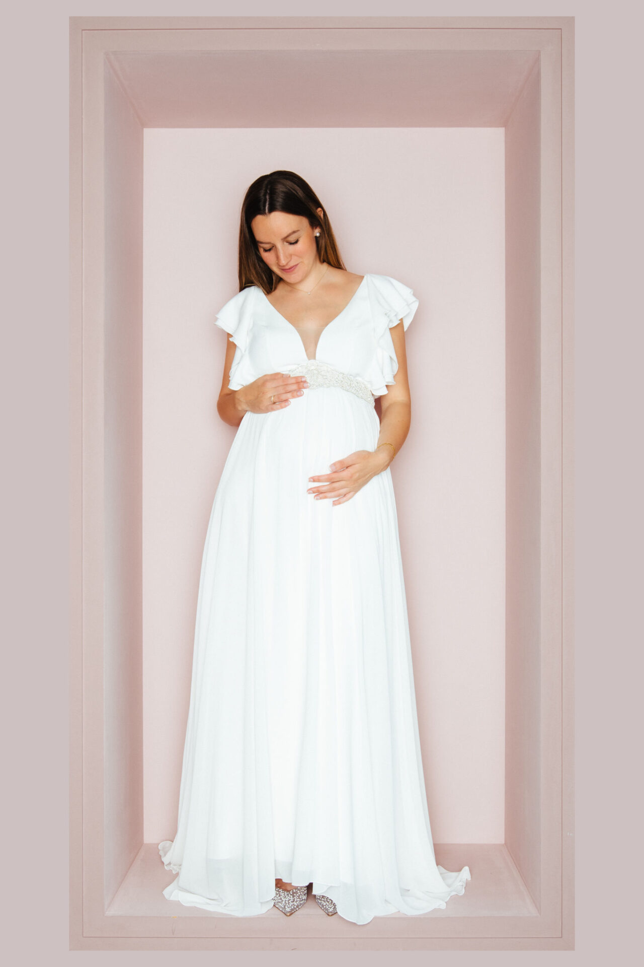 Schwangere Braut im Brautkleid mit tiefem V-Ausschnitt und Volantärmeln, die Hände am Babybauch