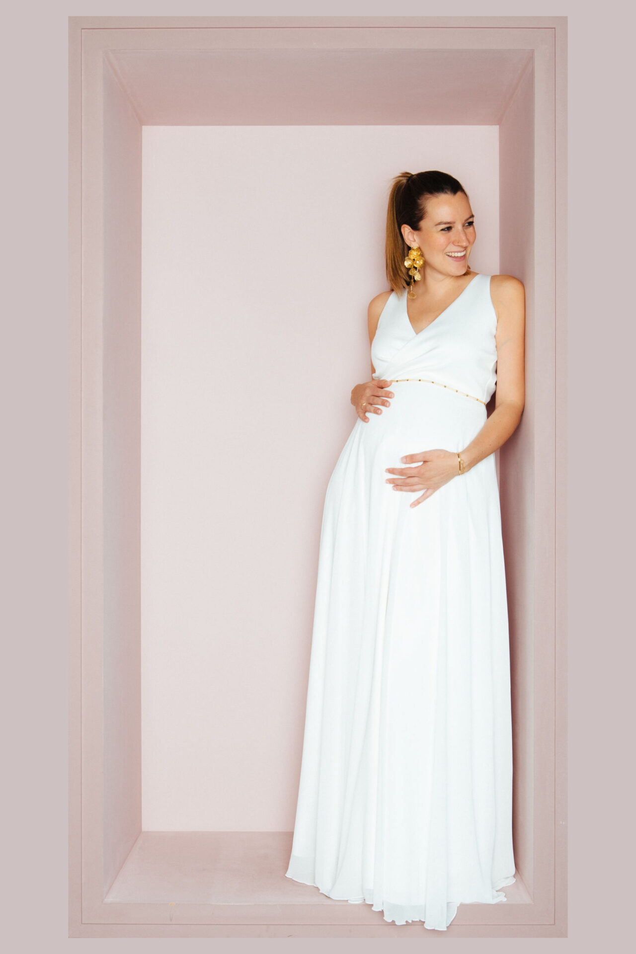 Schwangere Braut im ärmellosen Brautkleid mit V-Ausschnitt und weitem Chiffonrock, lächelnd an die Wand gelehnt