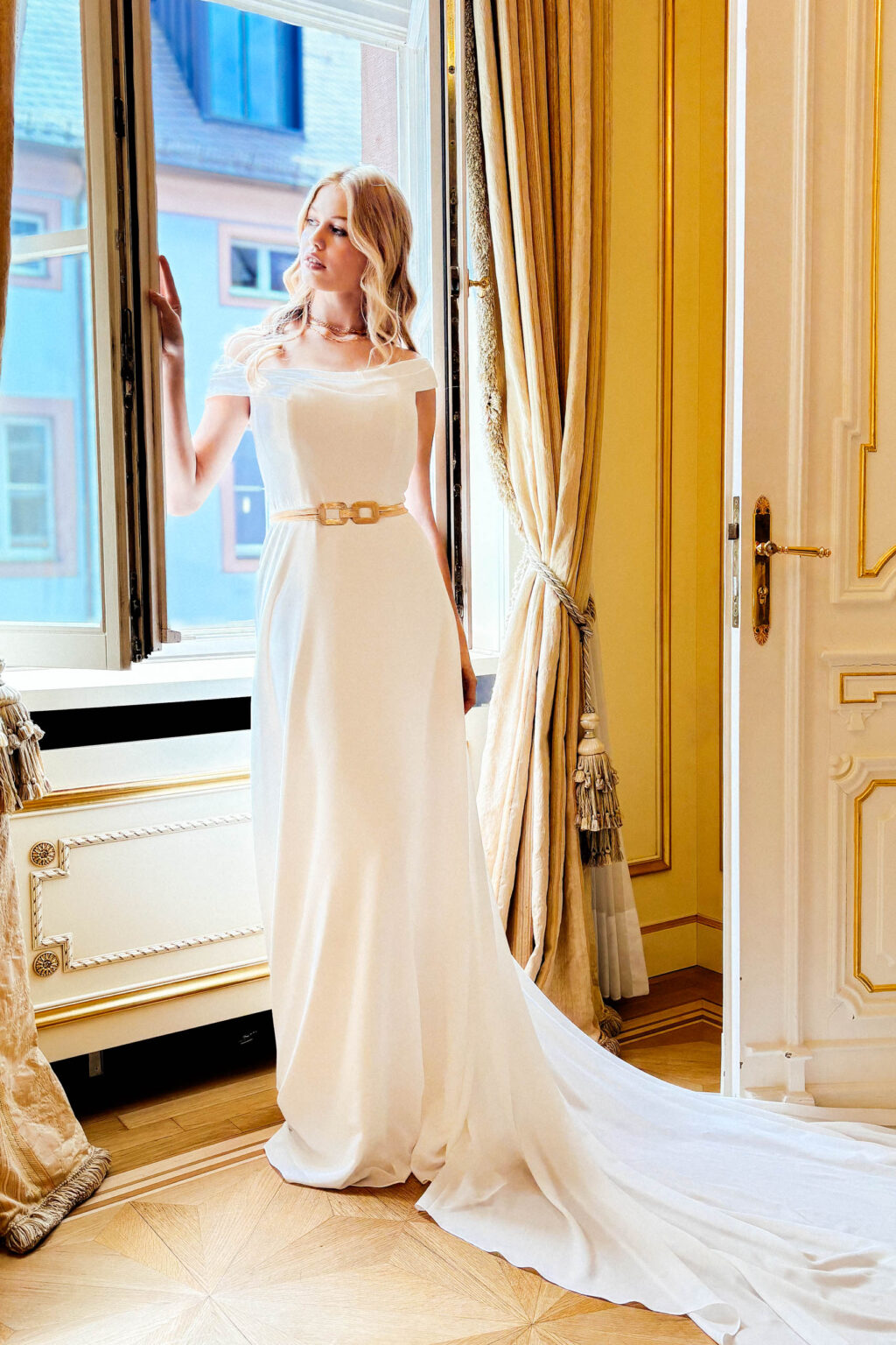 Langes Chiffon-Brautkleid KUESSdieCARLOW mit asymmetrischem Carmenausschnitt und goldenem Gürtel am Fenster